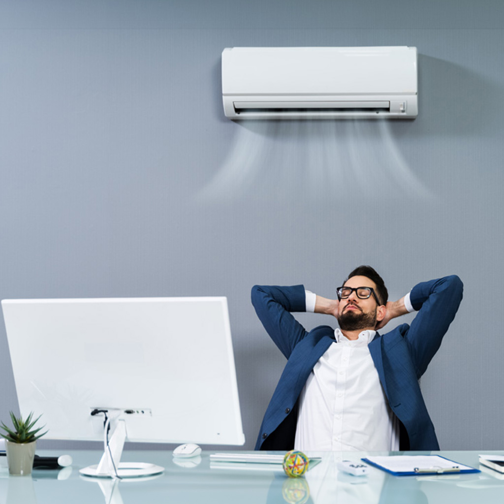 office worker leaning back at his desk with his hands behind his head enjoying the new office air conditioning unit that has been installed above him.